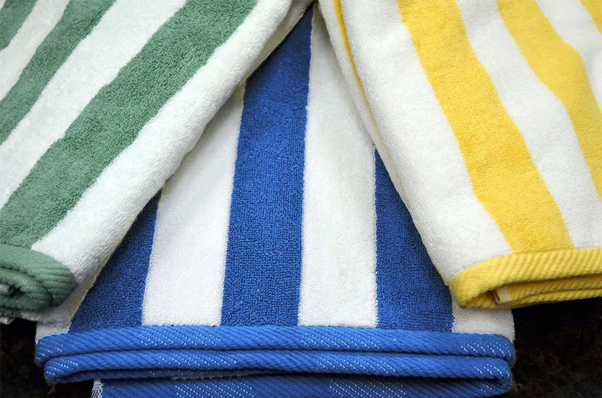 Three Ganesh Mills Cabana Stripe Beach Towels - Economy Vat Dyed are displayed folded and overlapping, featuring bold green/white, blue/white, and yellow/white stripes on a dark surface.