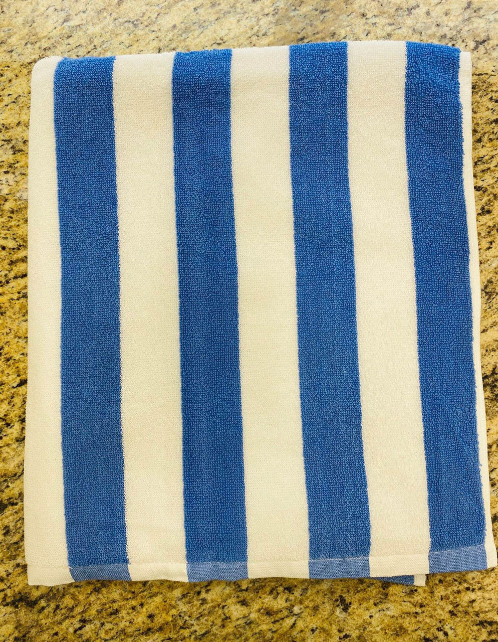 A folded Ganesh Mills Cabana Stripe Beach Towel – Economy Vat Dyed, featuring vertical blue and white stripes, rests on a tan speckled countertop.