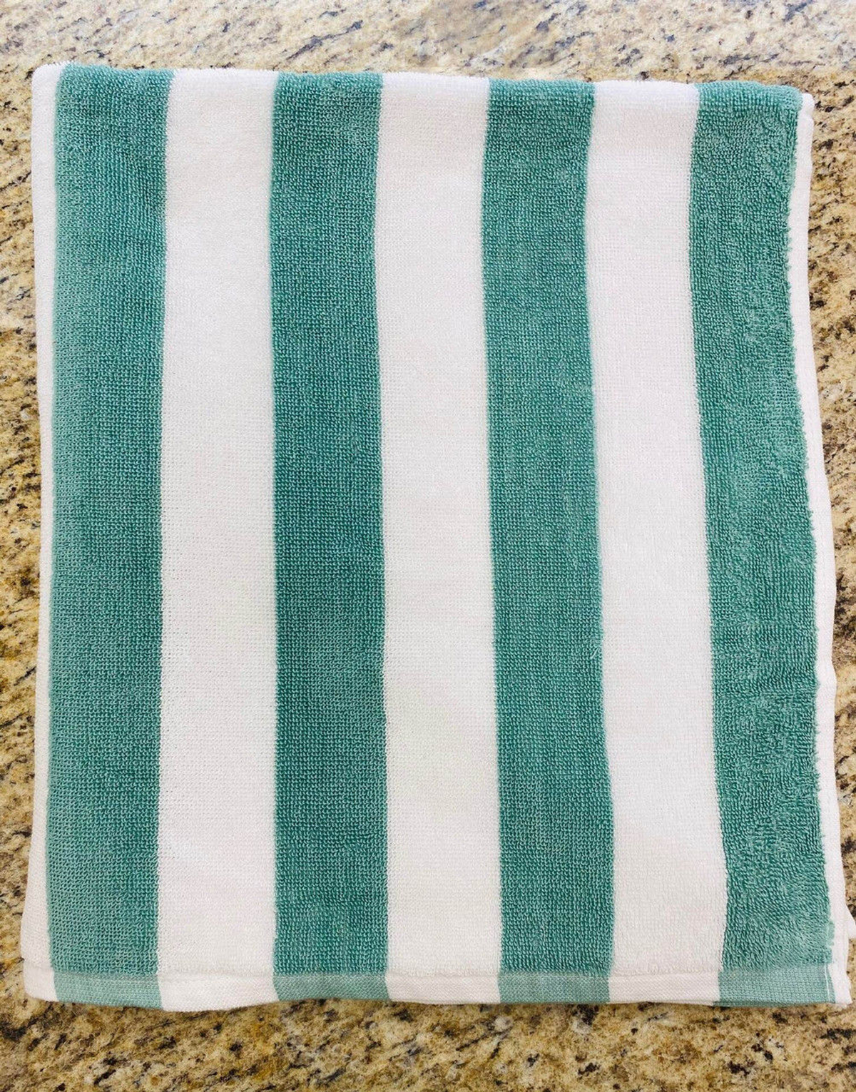 A folded Ganesh Mills Cabana Stripe Beach Towel - Economy Vat Dyed, featuring alternating teal and white stripes, rests on a speckled beige countertop.