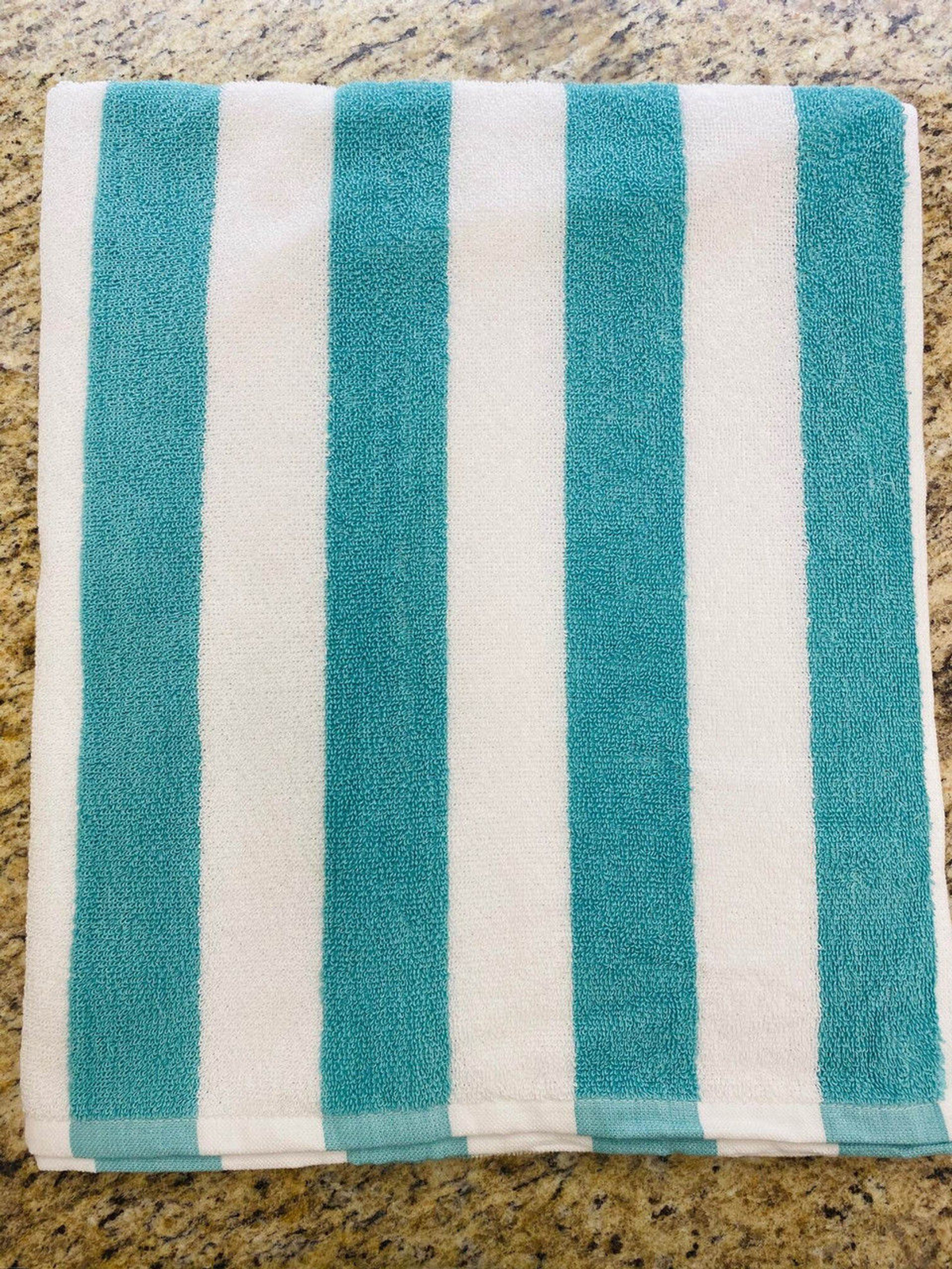A Ganesh Mills Cabana Stripe Beach Towel - Economy Vat Dyed, featuring vertical turquoise and white stripes, is folded and placed on a speckled beige countertop.