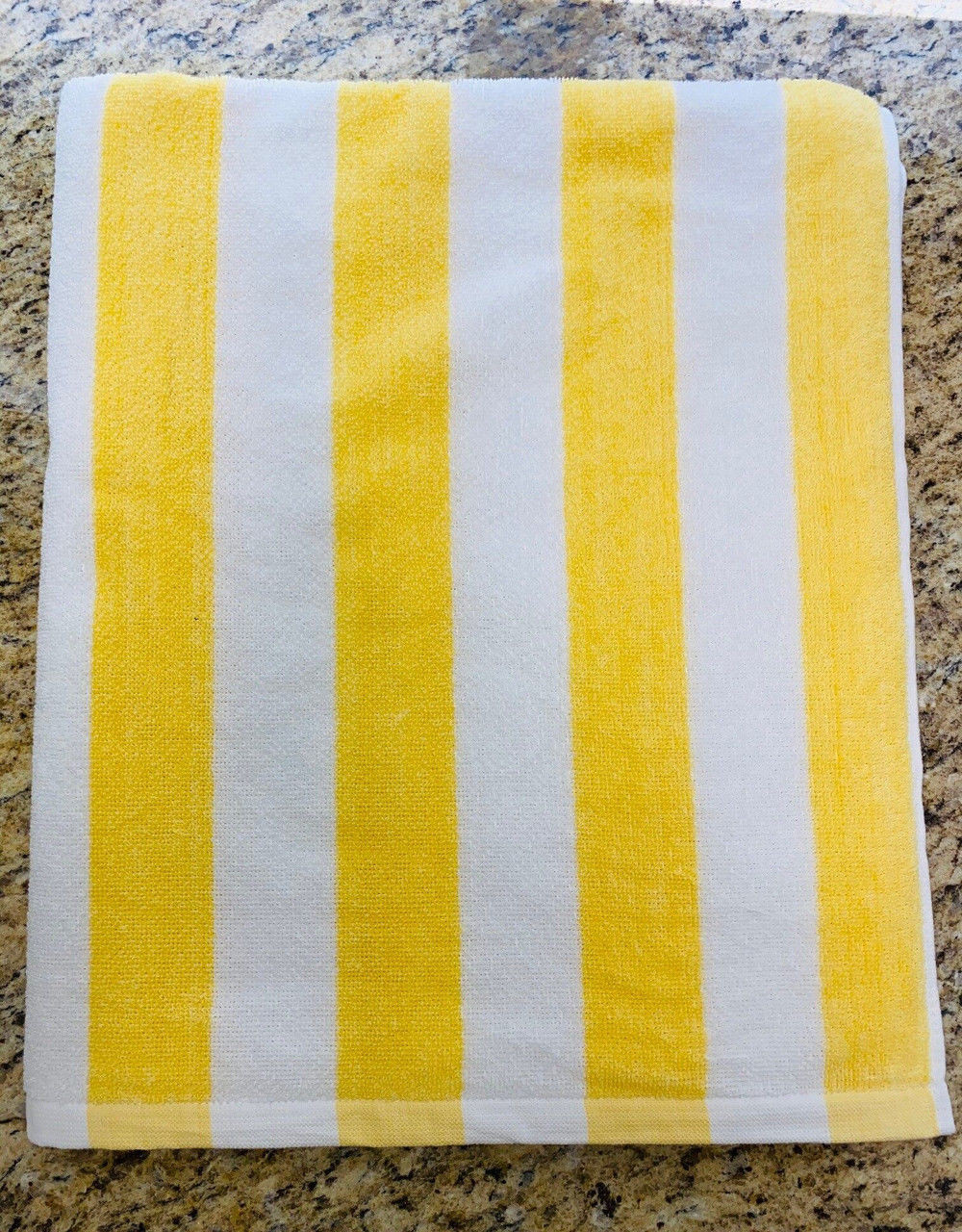 A Ganesh Mills Cabana Stripe Beach Towel - Economy Vat Dyed, featuring alternating yellow and white stripes, is folded on a speckled granite countertop.