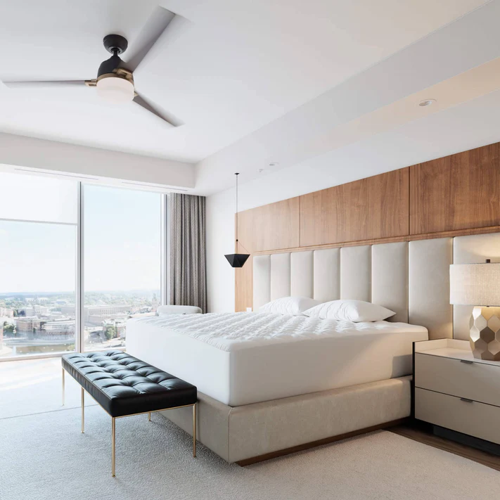 A modern bedroom with a large bed featuring the Bargoose Deluxe Microfiber Waterproof Quilted Mattress Pad (16" fitted skirt), tufted headboard, black bench, nightstands, lamps, and floor-to-ceiling windows with city views. Light walls and wood accents add warmth.