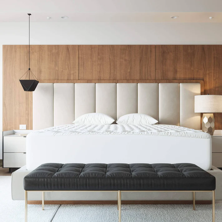 A modern bedroom with a large bed featuring a tall, tufted beige headboard, white bedding over the Bargoose Deluxe Microfiber Waterproof Quilted Mattress Pad, two white pillows, a black pendant light on the left, beige table lamp on the right, and a black bench at the foot.