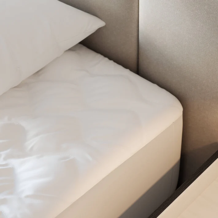 Close-up of a neatly made bed featuring the Bargoose Deluxe Microfiber Waterproof Quilted Mattress Pad with a 16" fitted skirt, and pillow, set by a gray upholstered headboard and dark bed frame in soft natural light.