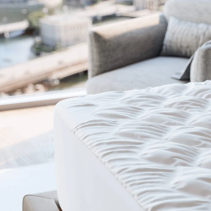 A close-up of the Bargoose Deluxe Microfiber Waterproof Quilted Mattress Pad with a 16" fitted skirt, featuring a white ruched finish on a bed near a window, with a grey armchair and cityscape in the background.