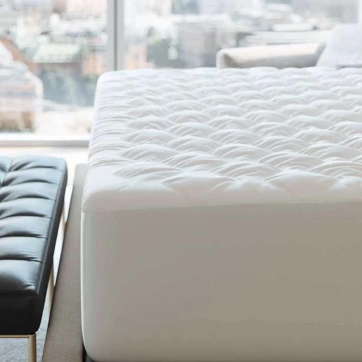 A close-up of the Bargoose Deluxe Microfiber Waterproof Quilted Mattress Pad with a 16" fitted skirt on a bed near a window, featuring a tufted black bench at the foot and a blurred cityscape outside.