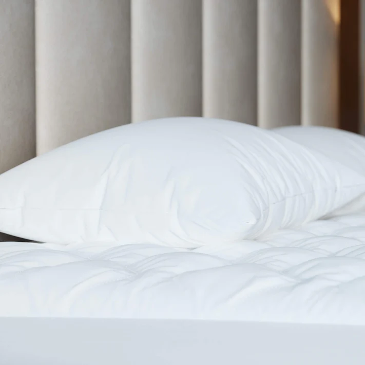 A white pillow and duvet rest neatly on a bed topped with the Bargoose Deluxe Microfiber Waterproof Quilted Mattress Pad with a 16" fitted skirt, while a padded, light beige headboard completes the inviting, minimal scene.