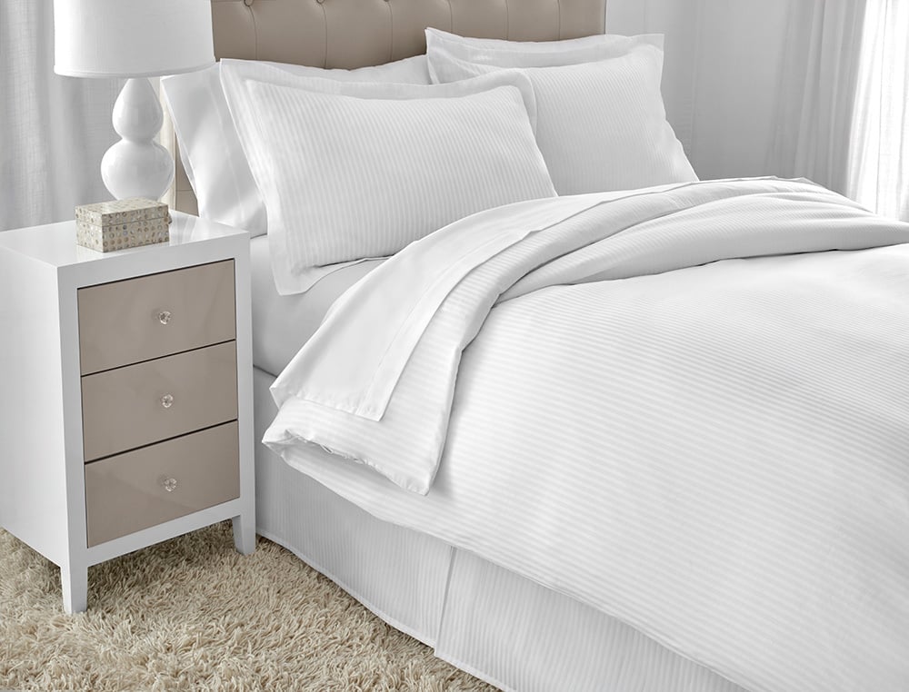 A neatly made bed features the WestPoint Home Five Star Hotel Collection DryFast 300 Duvet Cover and pillows, next to a white three-drawer nightstand with a lamp and tissue box, set on a beige shaggy rug in a bright room.