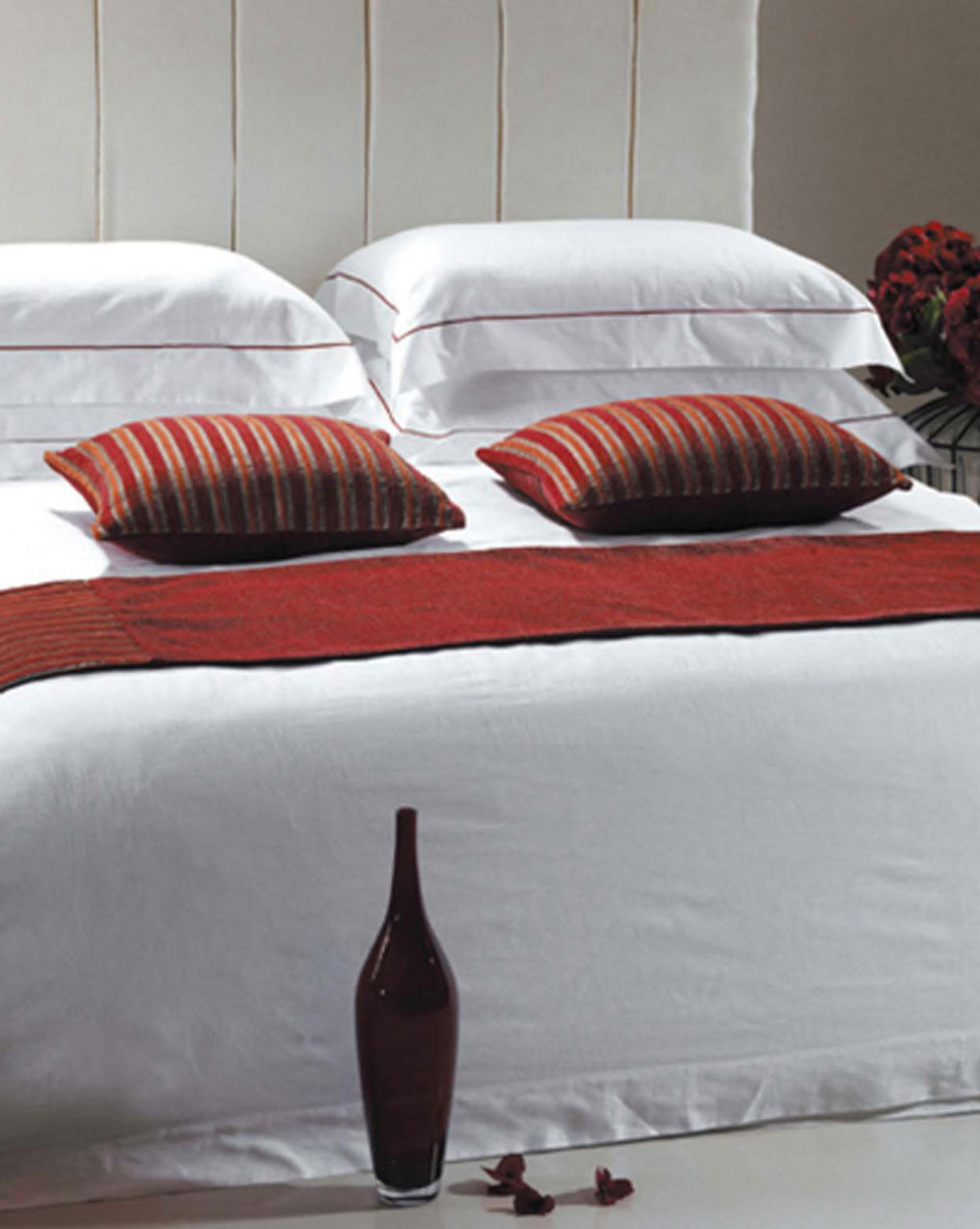 A bed features a Ganesh Mills Duvet Cover - Envelop Style with 15 Pocket Cotton/Poly Plain T250 Mercerized in white, two red-striped accent pillows, a red runner, a matching bottle with petals at the foot, and flowers on the right nightstand.
