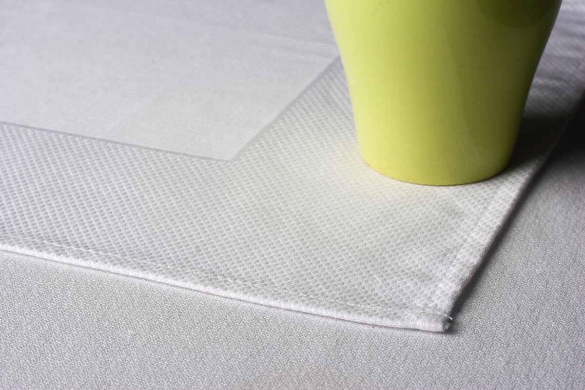 A light green cup sits on the corner of a textured white placemat atop the Ganesh Mills Tablecloth - 4 Sided Hemmed White Bird Eye Banquet Table Linen.