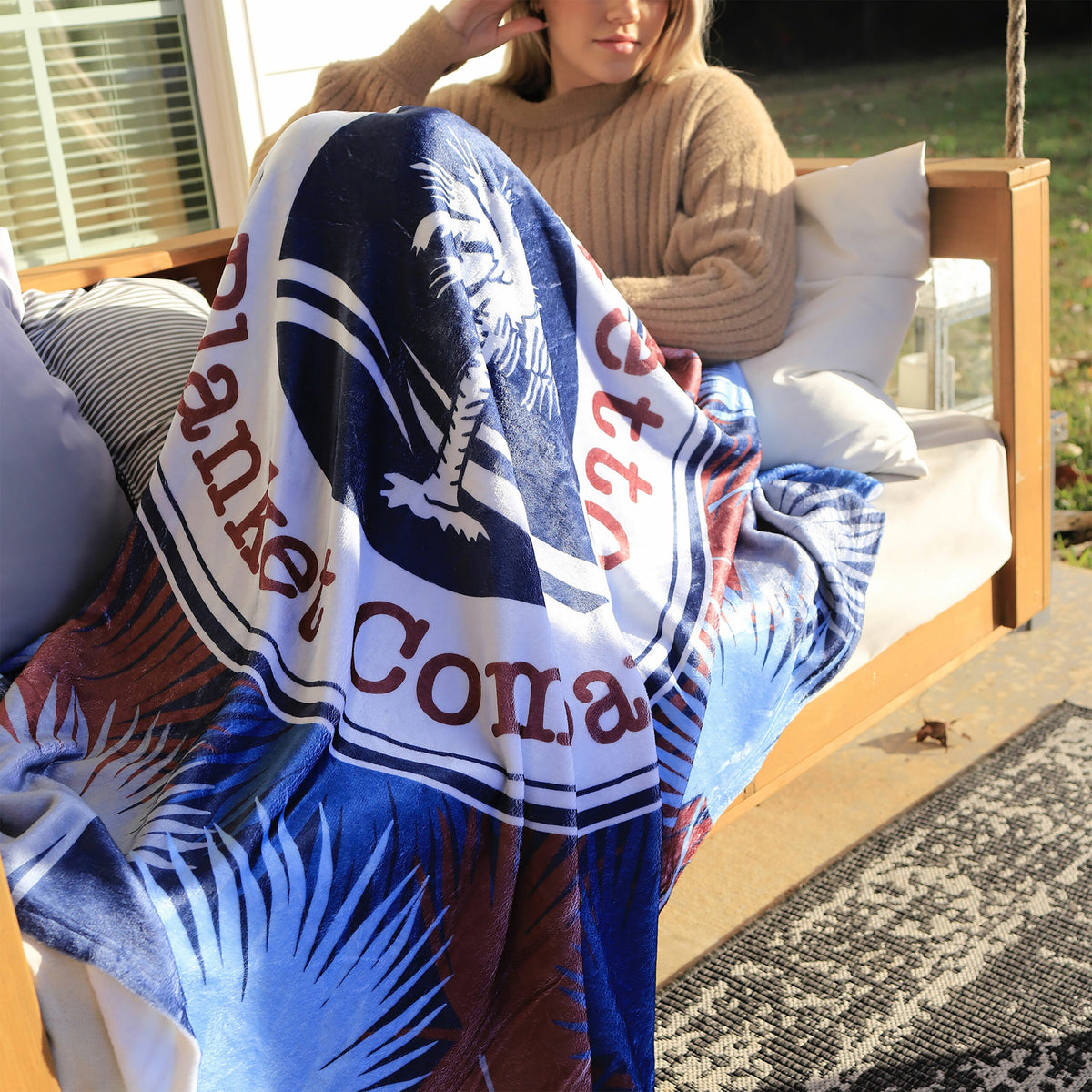 A person in a tan sweater sits on an outdoor swing with a Pro Vision Plush Throw by Pro Towels, featuring a bold blue, white, and red palmetto tree design and partially visible text. Sunlight warms the porch.