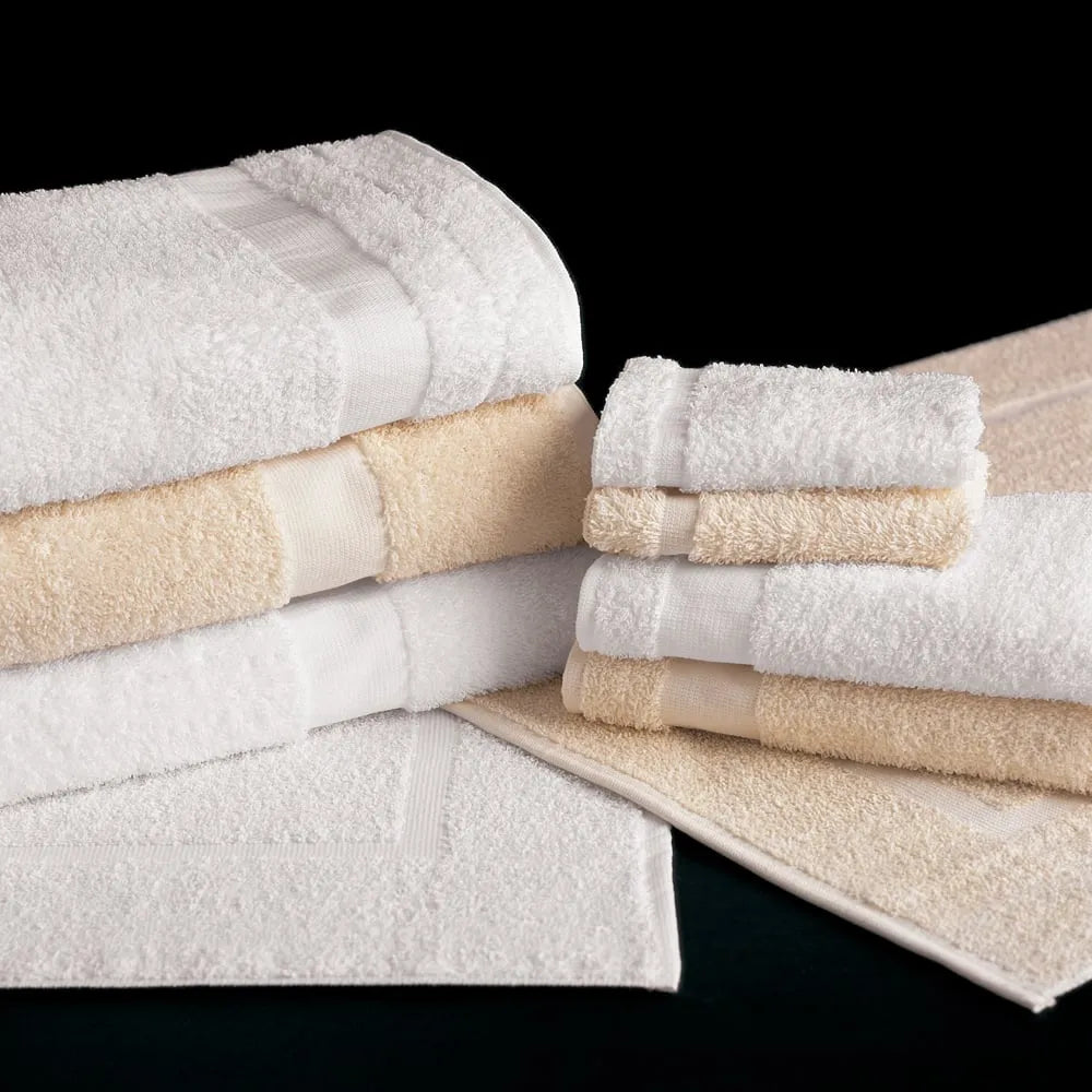 A set of WestPoint Home Martex Cam Towels, featuring white and beige cotton bath towels, hand towels, and washcloths, neatly stacked on a black surface with two bath mats partially visible underneath.