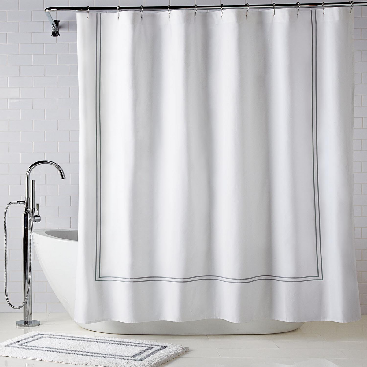 A white Supima cotton shower curtain with a thin gray border hangs above a freestanding tub. In front lies the Tufted Cut Bath Rug from the Gryphon Collection by WestPoint Home, on classic white subway tiles.