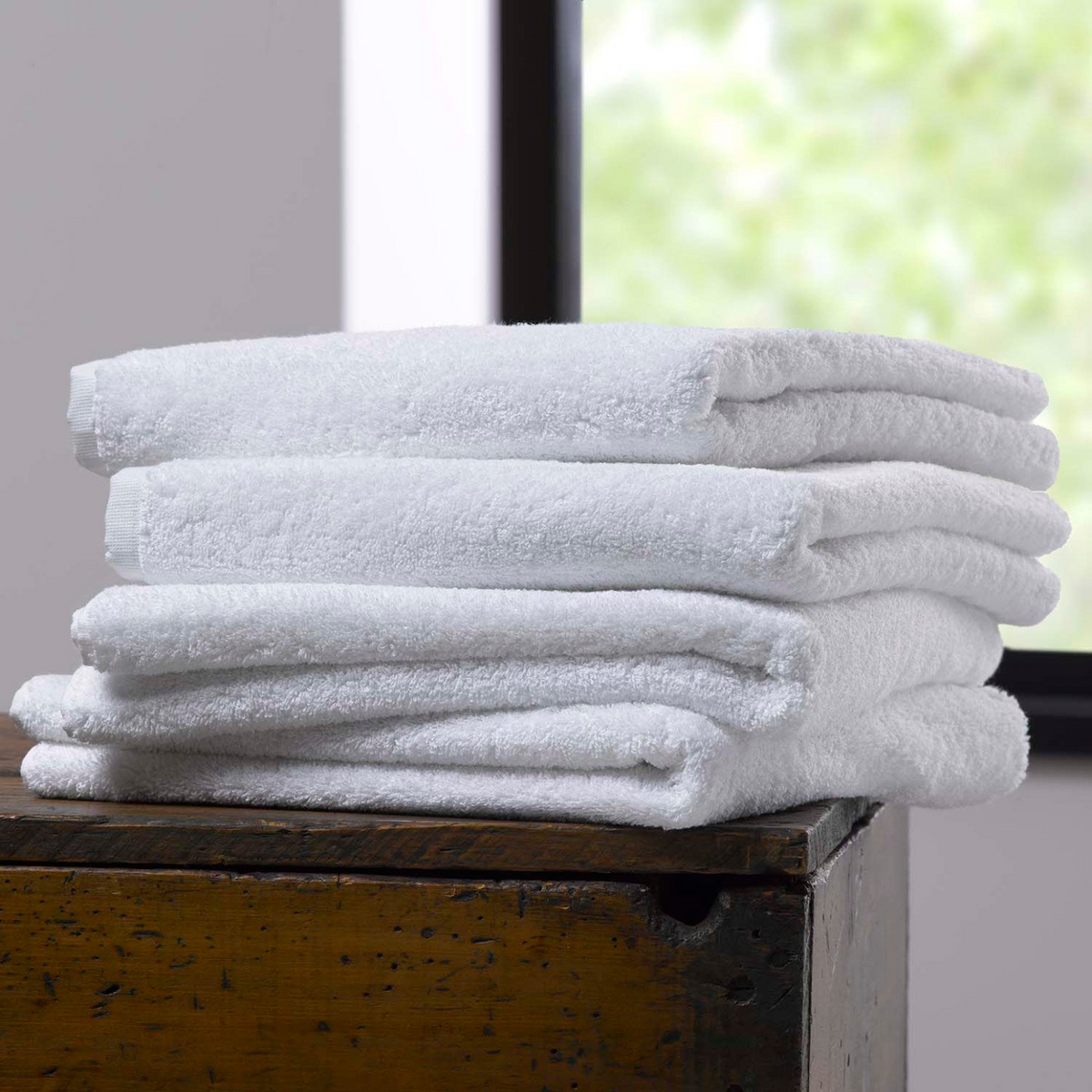 A neatly folded stack of WestPoint Homes Martex Green Dobby Towels, crafted from earth-friendly fabrics, rests on a wooden surface near a sunlit window with lush greenery outside.