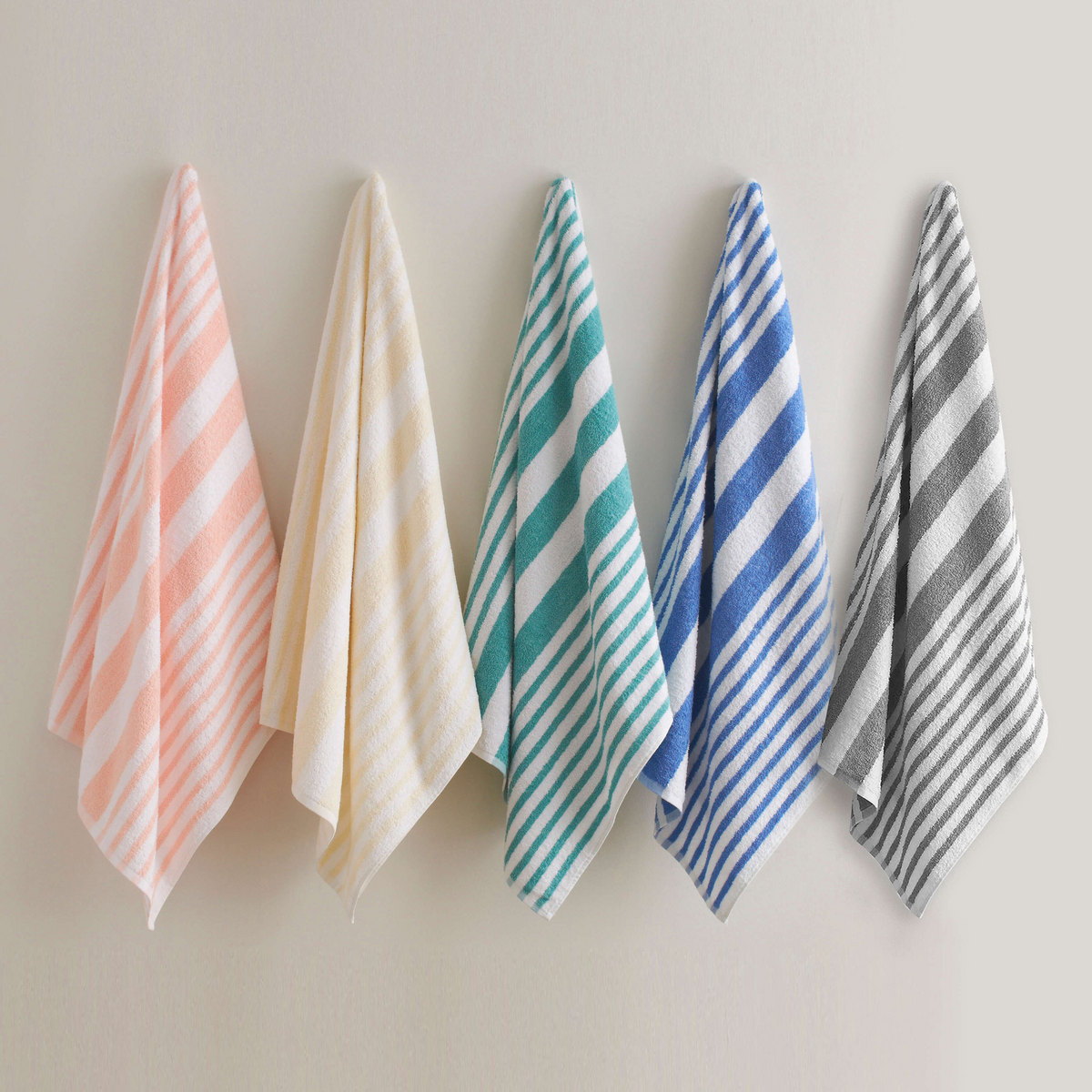 Five Martex Tropical Stripe pool towels by WestPoint Home in peach, yellow, green, blue, and gray hang side by side on a light wall—ideal for wholesale orders or adding vibrant color to any space.