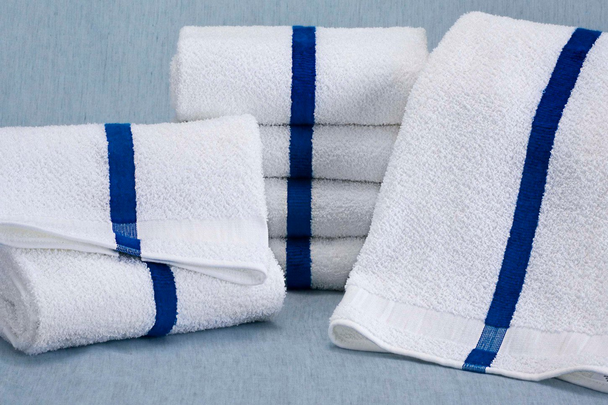 The Martex Cabana Collection Pool Towel by WestPoint Home features a white design with a single vertical blue stripe, neatly folded and stacked on a light blue surface—ideal for hospitality use or poolside relaxation.