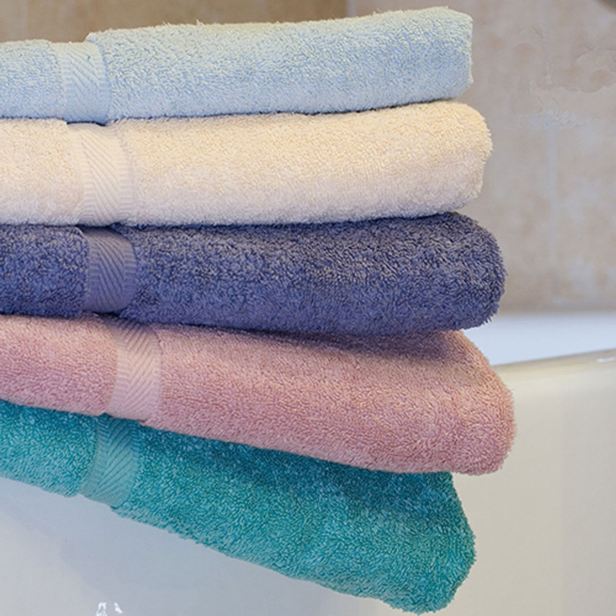 A stack of five Ganesh Mills Oxford Imperiale Color Bath Sheets in light blue, cream, navy blue, mauve, and teal is neatly folded by a white bathtub with a tiled wall in the background.