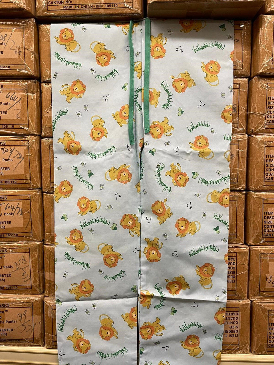 A white curtain with a playful yellow lion and green grass print—evoking KSE’s Lazy Lion Pediatric PJ Pants—hangs before stacked brown Girls Panty boxes in a warehouse.