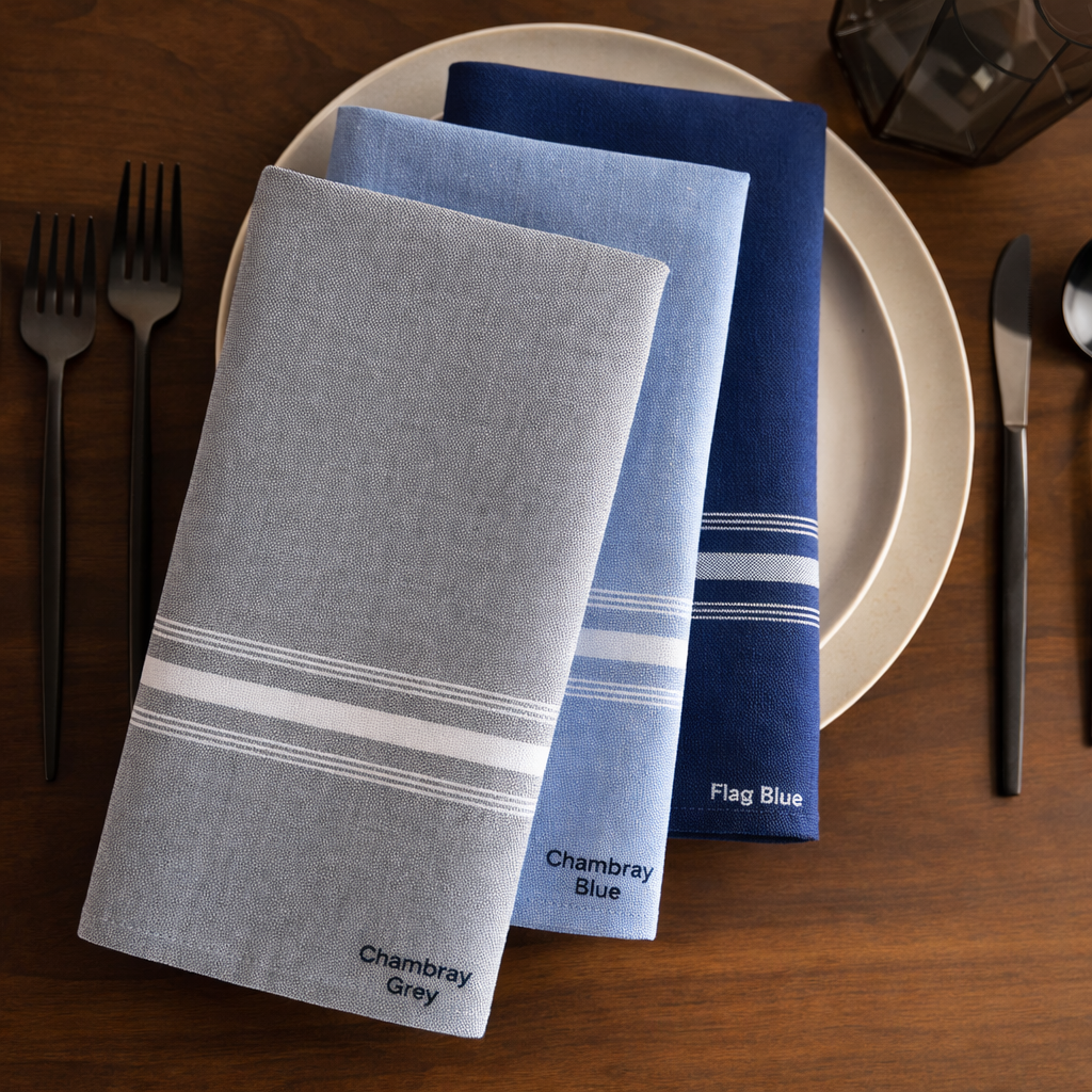 Three folded PREMIER BISTRO TABLE LINEN napkins by 1Concier—Chambray Grey, Chambray Blue, and Flag Blue—each with a yarn-dyed stripe, are arranged on a plate atop a wooden table with black cutlery and a geometric candle holder. Ideal for commercial use.