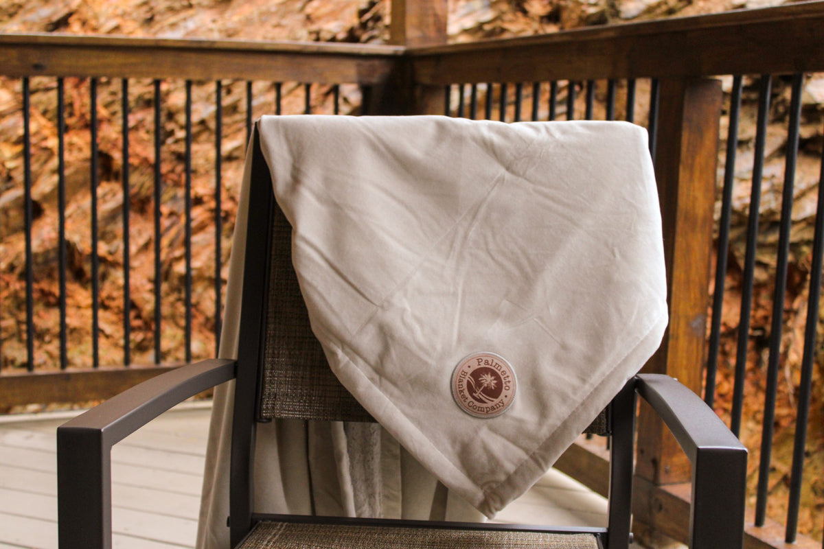 A Pro Towels Opulence Throw featuring custom embroidery and a brown circular logo is draped over a patio chair on a wooden deck, with railing and rocks visible in the background.