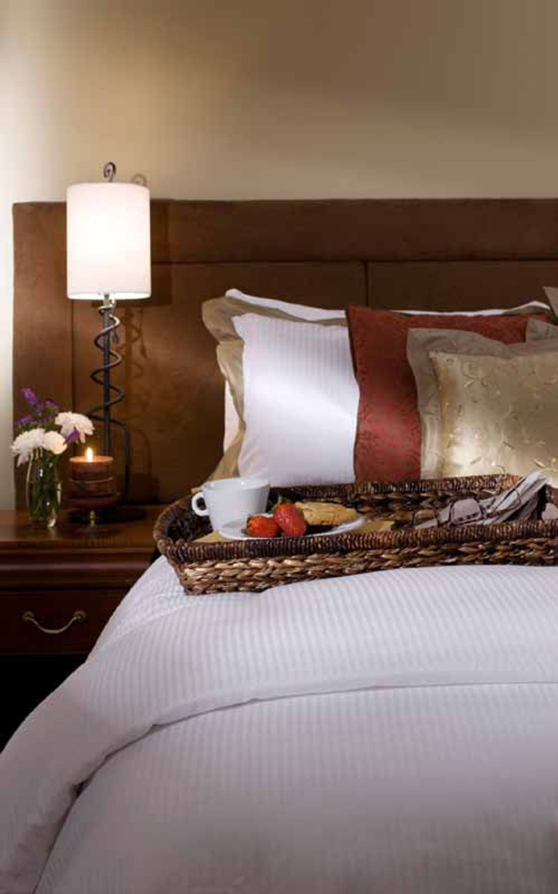 A neatly made bed with white linens and a Ganesh Mills Pillow Sham - 5mm Tone On Tone Stripe T250 Mercerized holds a wicker tray with breakfast, strawberries, and a mug. A bedside table displays a lamp, candle, and small vase of flowers.