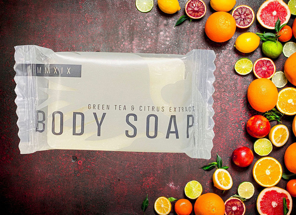 A Ganesh Mills Soap (Rectangle Shape) - Candy Wrap Embossed bar in clear packaging sits on a red surface, surrounded by assorted oranges, limes, and lemons—some whole, some sliced.