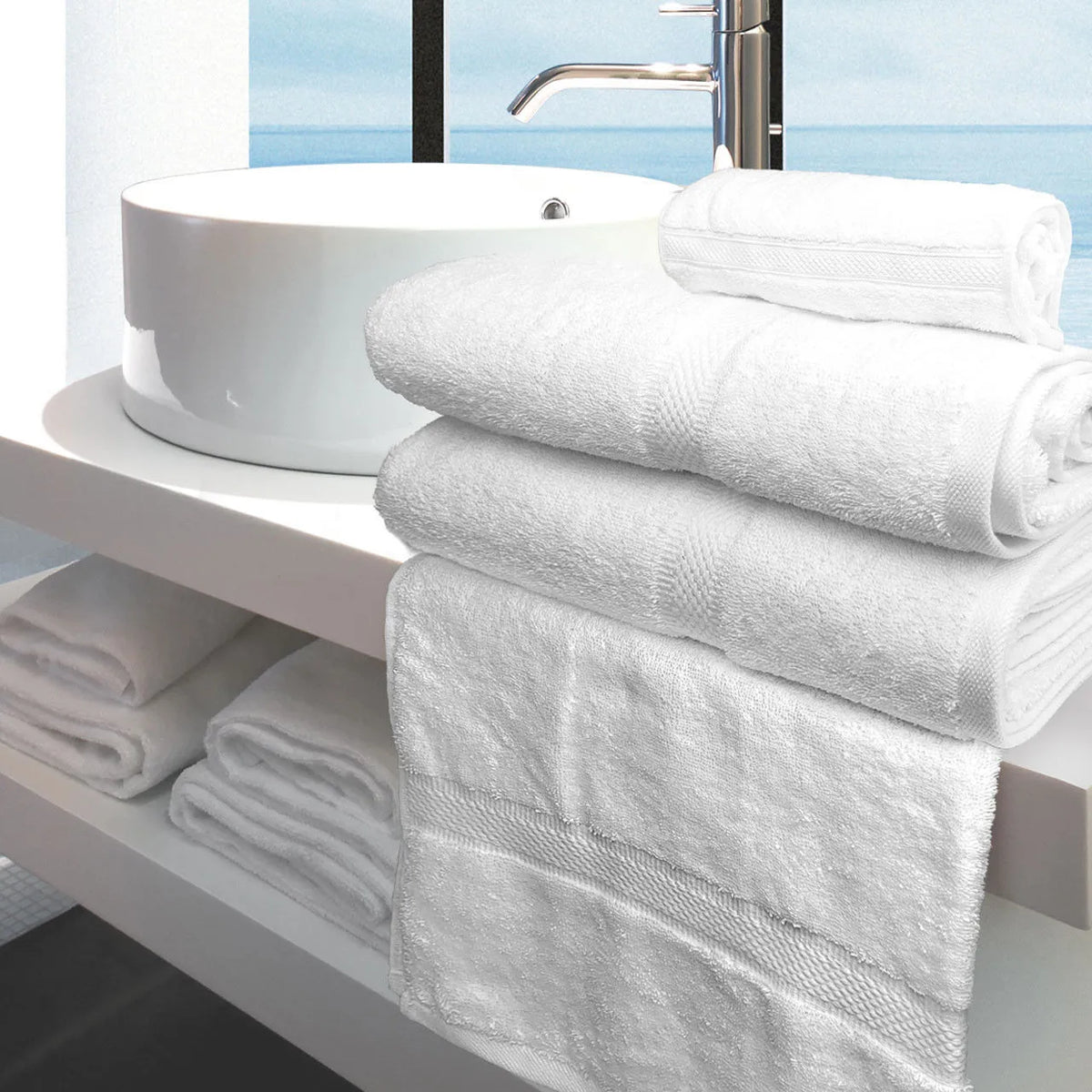 A stack of Ganesh Mills Washcloth - White Oxford Imperiale Max Towel Collection sits on a modern white bathroom counter with more towels and washcloths on a shelf below, and a window overlooking water in the background.
