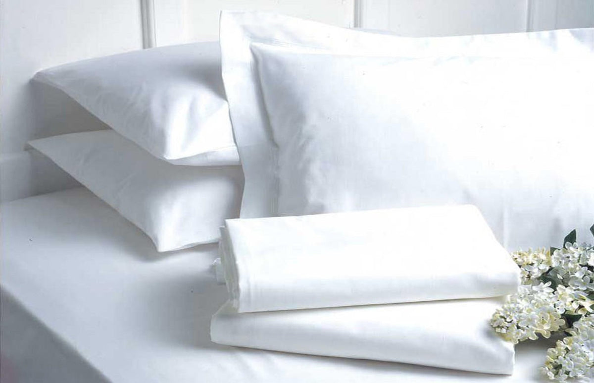 Two white pillows sit on a neatly made bed with a white sheet, beside a folded Ganesh Mills Pillow Case - Oxford Micro Superblend Bed Linen Antistatic and a small bunch of white flowers, creating a clean and fresh look.