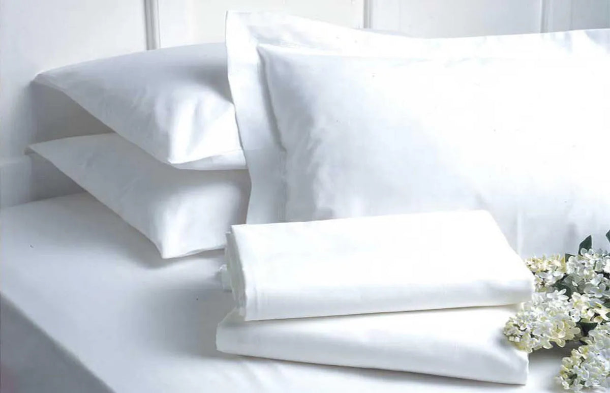 A neatly stacked set of Ganesh Mills Oxford Micro Superblend Bed Linen Flat Sheets with 2" top hem and 1" bottom hem, plus pillowcases, rests on a bed near white flowers against a minimal white background.