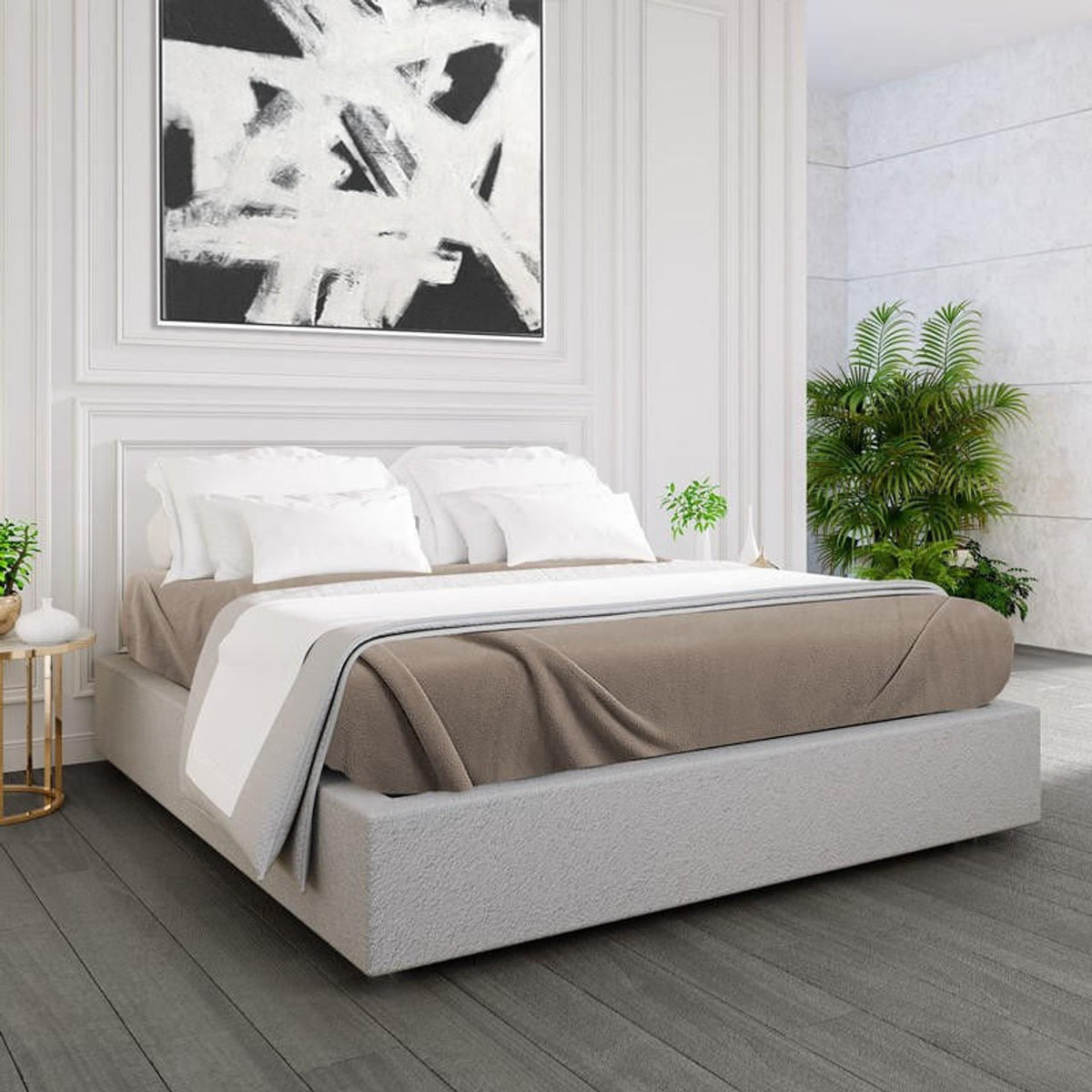 A modern bedroom features a low upholstered bed with white bedding and a beige Ganesh Mills Oxford Polar Fleece Blanket. A bold black-and-white abstract painting hangs above, accented by gold side tables and green potted plants in the corner.