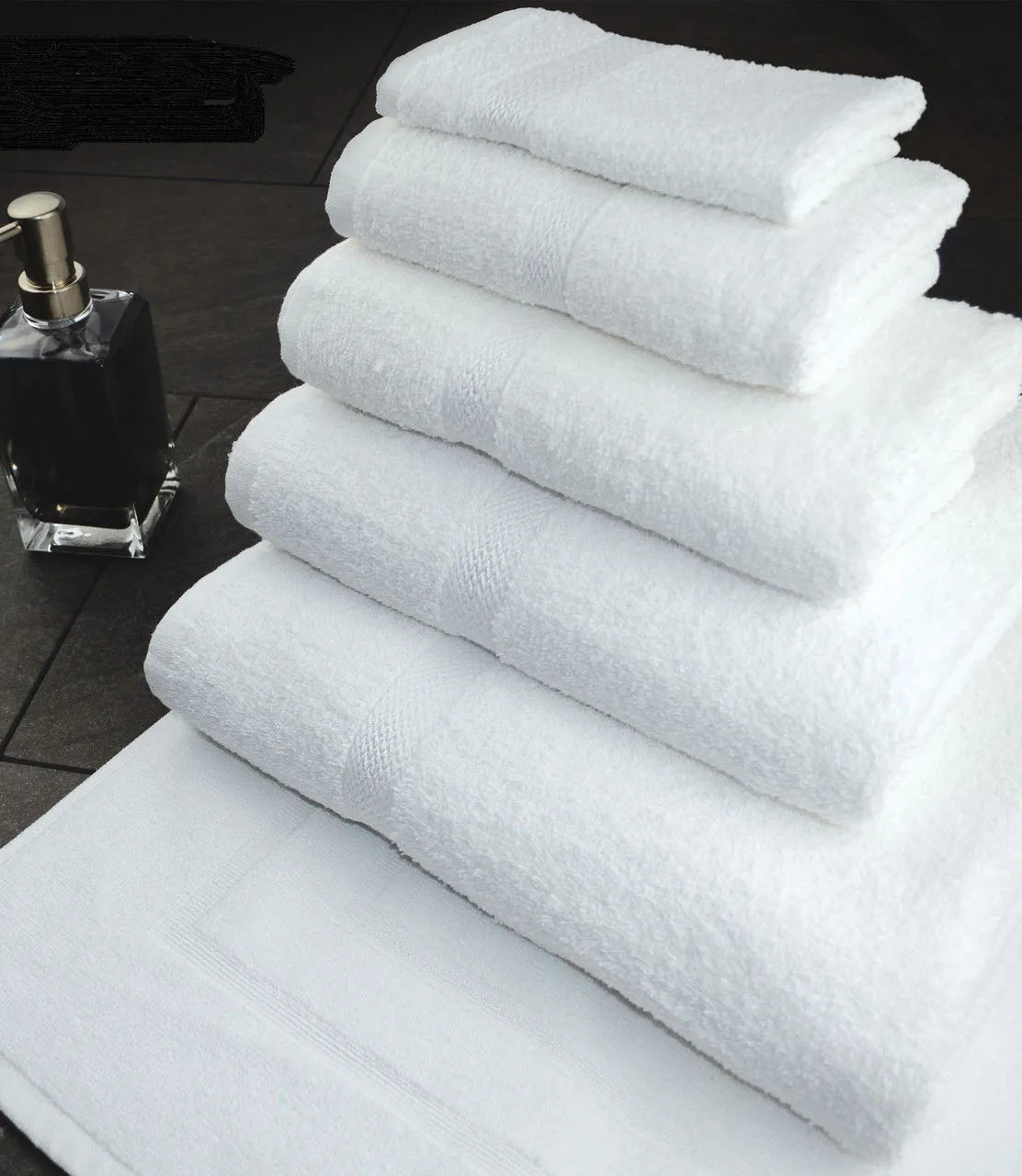 A stack of Ganesh Mills Bath Sheet / Pool Towels – White Oxford Nuvola is arranged by size next to a clear square bottle with a metallic pump, all placed on a dark tiled surface.