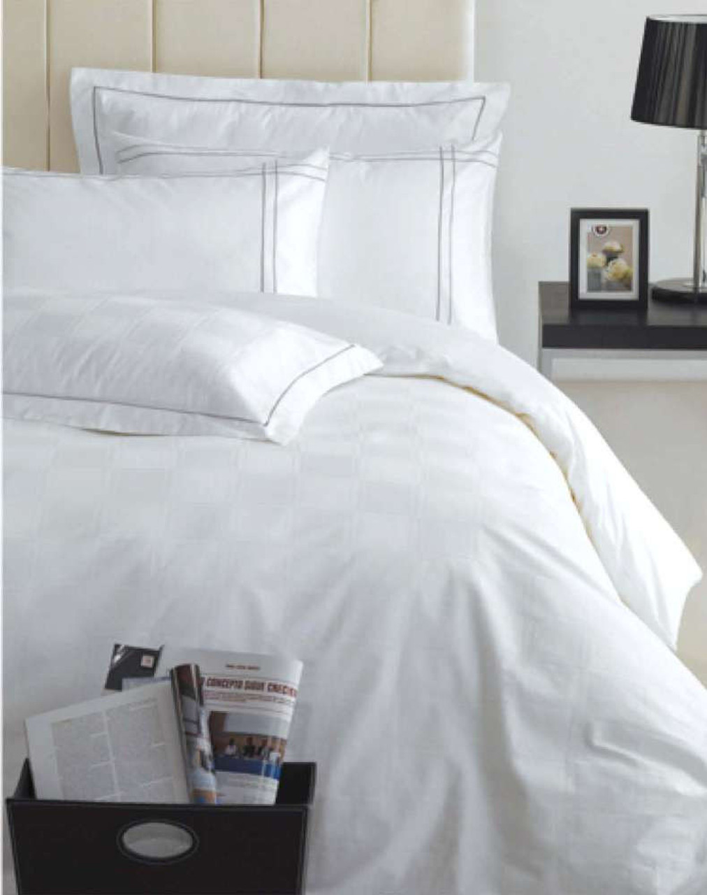 A neatly made bed with Ganesh Mills Oxford Pillow with Microgel and white bedding sits beside a nightstand holding a lamp and framed photo. A storage box at the foot of the bed holds a book and newspapers.
