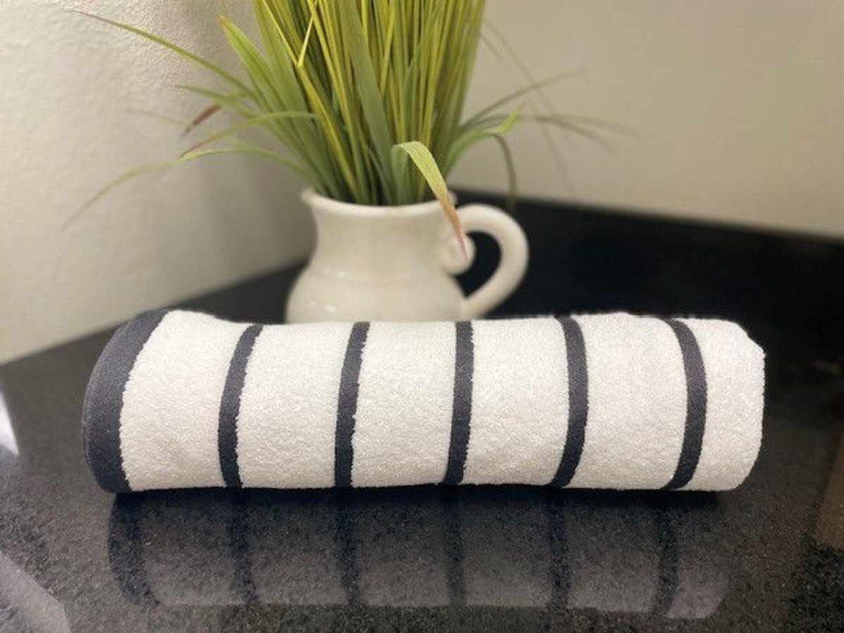 The Ganesh Mills Oxford Premium Horizontal Stripe Pool Towel in black lies on a dark countertop, with a white pitcher of green artificial grass bringing a fresh accent to the scene.
