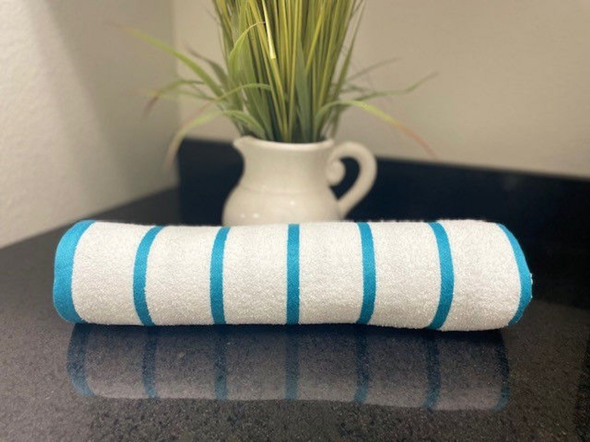 The Oxford Premium Horizontal Stripe Pool Towel by Ganesh Mills, featuring turquoise stripes, is rolled up on a black countertop with a white pitcher of green grass in the background.