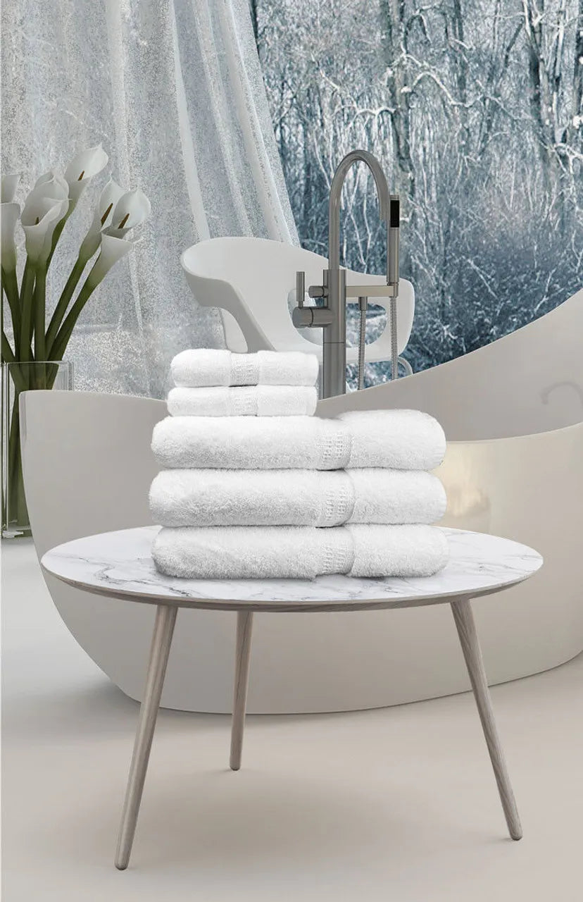 A stack of white Ganesh Mills Oxford Reserve Towels and a Bath Mat rest on a marble table in a modern bathroom, with snowy forest views framed by large windows.