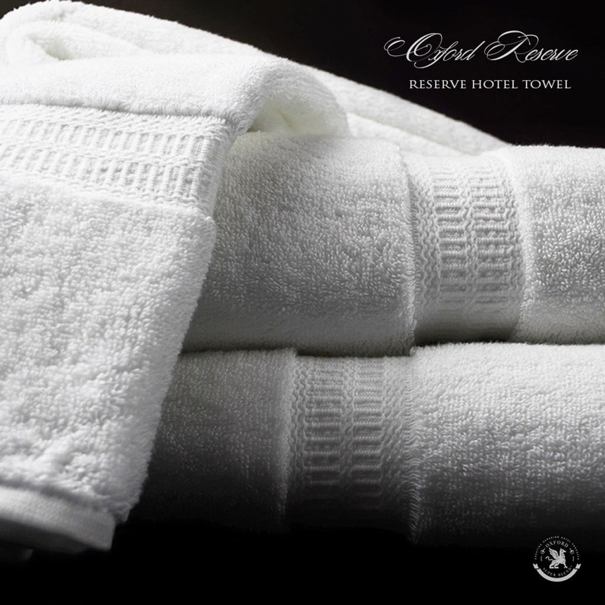 Two neatly folded, plush white washcloths from the Ganesh Mills Oxford Reserve Towel Collection are stacked against a dark background, showing their soft texture. "Washcloth - White Oxford Reserve Towel" appears in the top right corner.