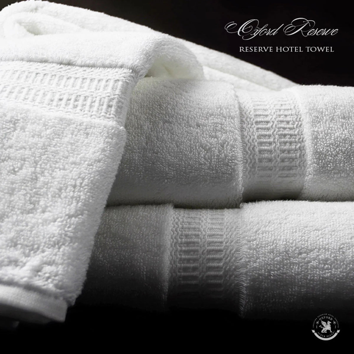 A neatly folded, white Bath Mat from the Oxford Reserve Towel Collection by Ganesh Mills sits on a dark surface, featuring ribbed detailing and a small logo in the lower right corner, adding elegance to any space.