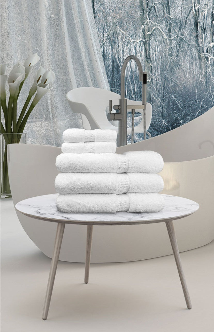 A stack of Ganesh Mills White Oxford Reserve washcloths sits on a round marble table in a modern bathroom with a freestanding tub, flowers in a vase, and large windows displaying a snowy winter forest outside.