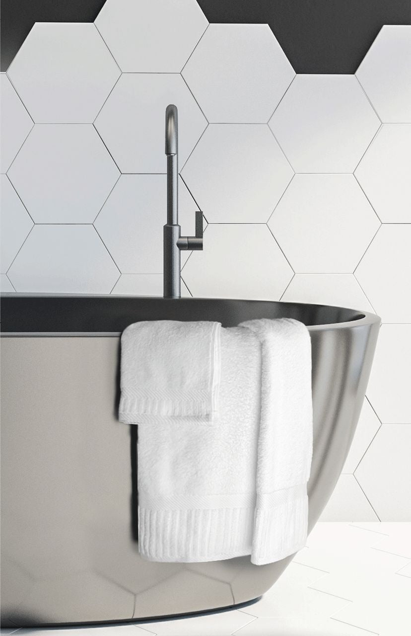 A modern bathroom with a large gray freestanding tub, sleek silver faucet, and the Ganesh Mills Washcloth - White Oxford Signature Towel draped over the edge. The hexagonal white tiled wall adds a luxe, hotel-inspired touch.