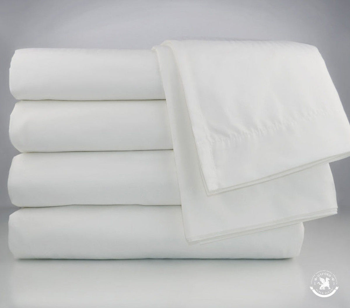 Four neatly folded Pillow Case 2 Hem - Oxford T180 Superblend Bed Linen by Ganesh Mills are stacked, with a fifth draped over the pile and crisp white oxford pillowcases, all set against a plain gray background.