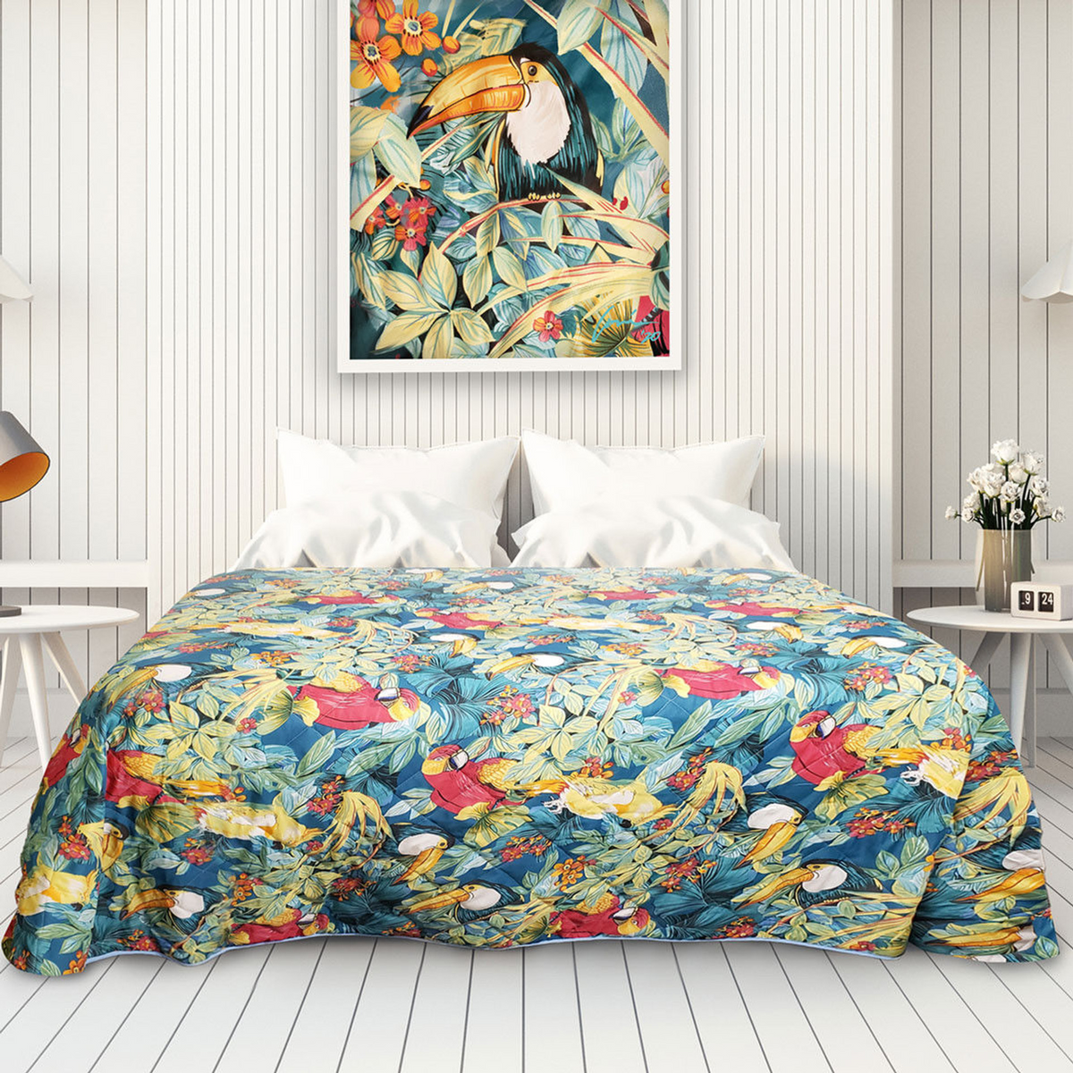 A Ganesh Mills Oxford Printed Bedspread with vibrant birds and leaves decorates the bed, complemented by white nightstands and lamps, while a framed toucan artwork hangs above in a bright, white-paneled room.