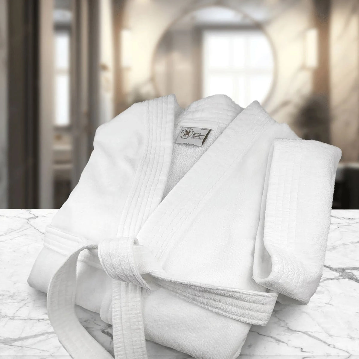 A Shawl Collar - White Oxford Velour Bathrobe by Ganesh Mills, neatly folded with a matching towel, rests on a marble surface. The softly blurred background reveals a bright room with large windows and neutral decor.