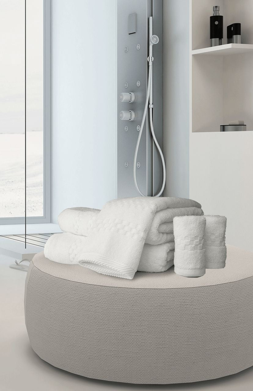 A modern bathroom features a round beige ottoman with luxury hotel towels and a Ganesh Mills Washcloth - White Oxford Viceroy Towels. A glass shower with a handheld showerhead and black bottles creates a serene atmosphere in the background.