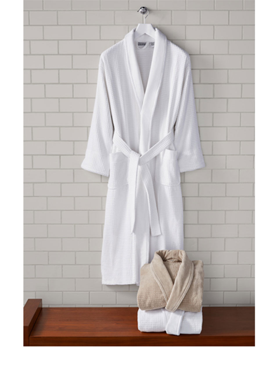 A Platinum Bathrobe by 1Concier hangs on a hook against a light gray tiled wall. On the wooden bench below, two folded 1Concier robes—one tan, one white—feature soft terry velour and are ideal for custom embroidery.