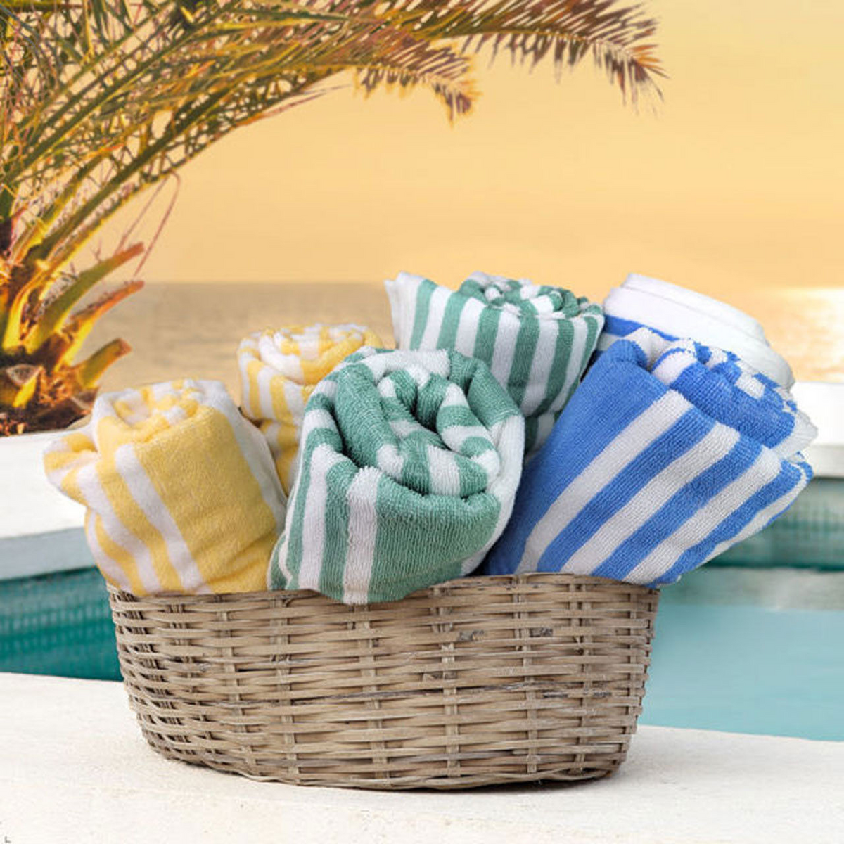 A wicker basket filled with Ganesh Mills’ Oxford Playa 2x2 Cabana Pool Towels in yellow, green, and blue sits poolside, framed by palm leaves and a sunset over the water.
