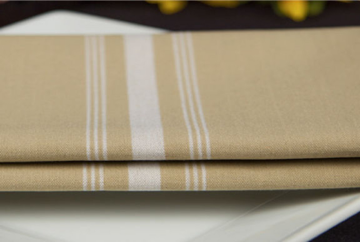 A folded beige Premier Bistro Napkin by 1Concier with white stripes rests on a white plate, set against a blurred background.