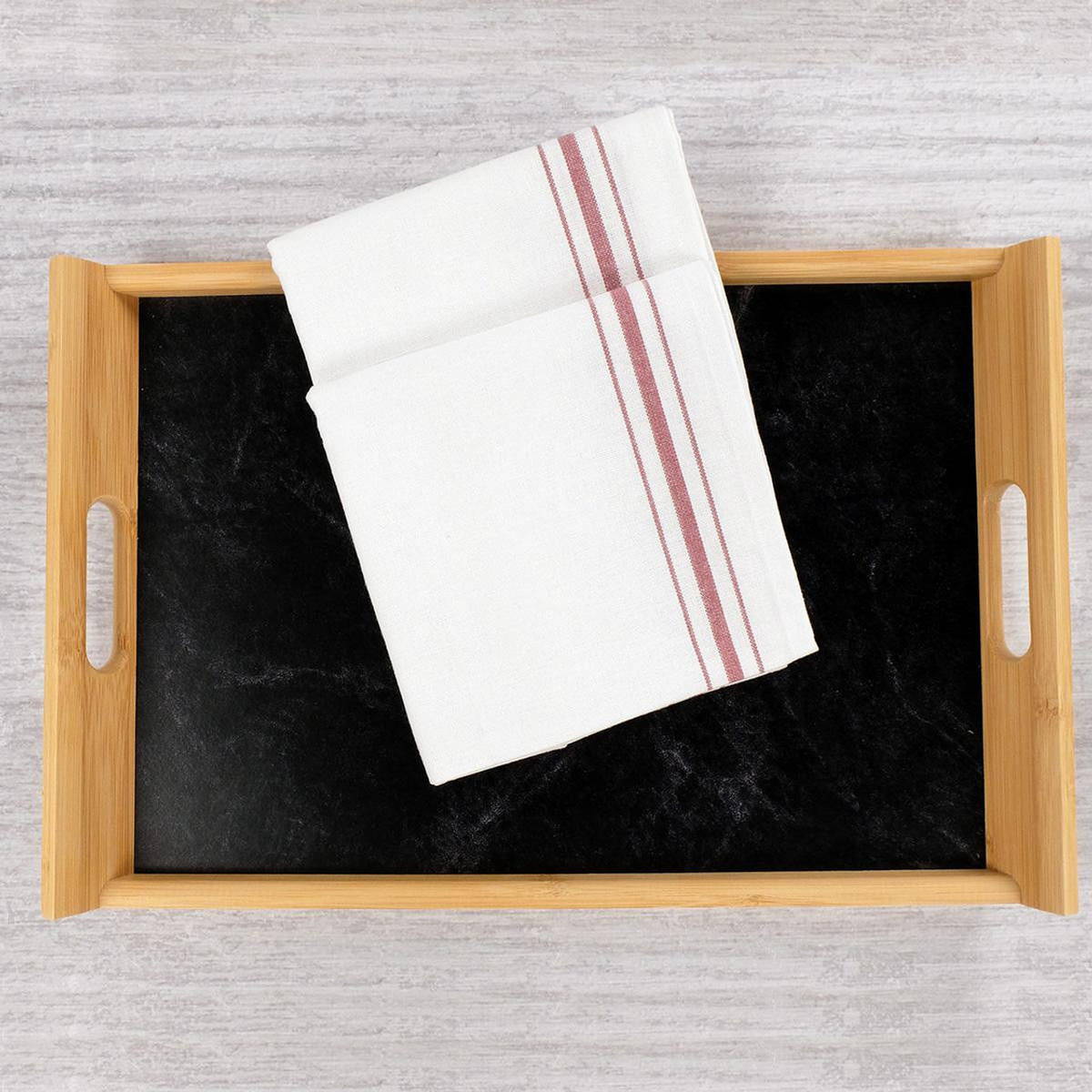 A wooden tray with handles holds a folded 1Concier Napkin Bistro—100% cotton, white with black stripe. The tray sits on a black surface, set against a subtly textured light gray background.