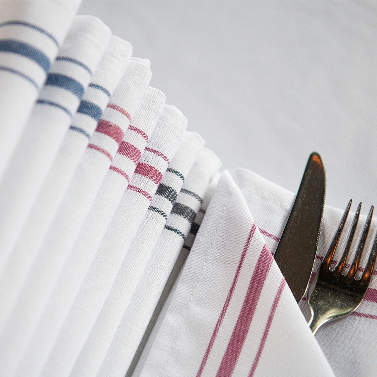 A close-up of neatly folded Napkin Bistro 100% Cotton White / Black Stripe by 1Concier, beside a knife and fork partially wrapped in one napkin, all set on a white tablecloth.