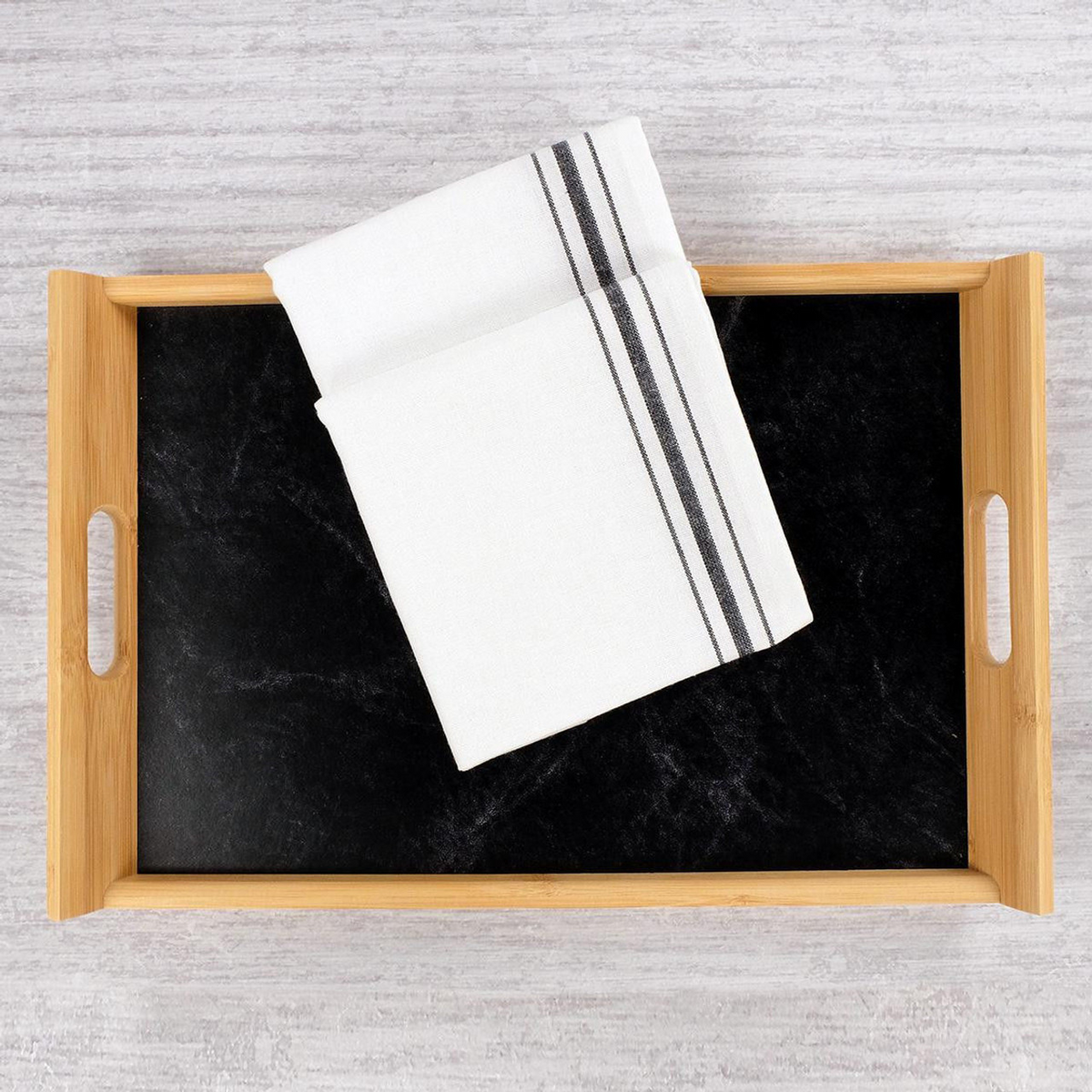 A wooden tray with a black base holds the Napkin Bistro 100% Cotton White / Black Stripe by 1Concier, featuring two black stripes along one edge, folded and placed on a light-textured surface.
