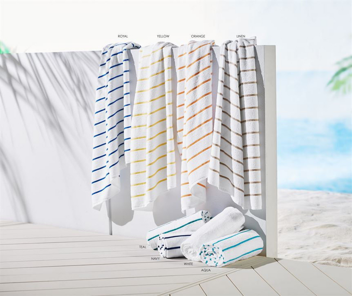 Five white 1Concier Pool Towel Ritz (20 lbs) with poly neon stripes in blue, yellow, orange, beige, and teal hang on a white partition above folded towels. Crafted from 100% cotton loops, they pop against a sandy beach and ocean backdrop.